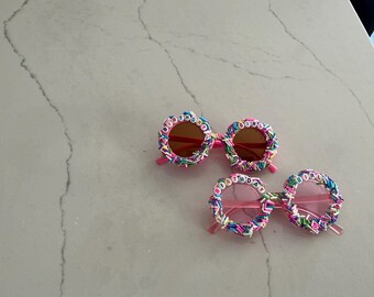 May include: Two pairs of pink sunglasses with brown lenses. The rims are decorated with colorful sprinkles and the word "BIRTHDAY" in letter beads. A fun accessory for a birthday celebration.