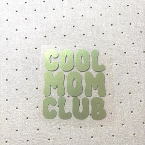 Könnte beinhalten: Ein hellgrüner Wärmeübertragungs-Vinyl mit den Worten "COOL MOM CLUB" in einer Bubble-Schrift. Der Text befindet sich auf einer transparenten Folie, vor einem weißen Stoff mit kleinen, gleichmäßig verteilten braunen Punkten.