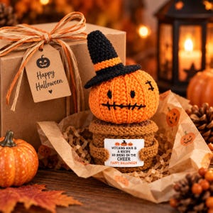 May include: A crocheted orange pumpkin wearing a black witch hat, sitting on a brown crocheted base. The pumpkin holds a white sign that reads: WITCHING HOUR IS A RECIPE SO BRING ON THE CHEER HAPPY HALLOWEEN. It is placed in a gift box with a tag.