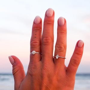 May include: Two delicate rings with small, light-coloured gemstones are displayed on a hand. The rings have thin, silver bands. The background shows a beach scene with a soft, colourful sunset.