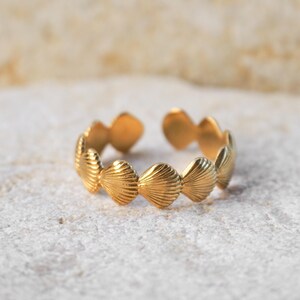 May include: A gold ring with a seashell design. The ring is open at the top and is resting on a light-coloured, textured surface. The seashells have detailed ridges. The ring is likely made of gold-plated metal.