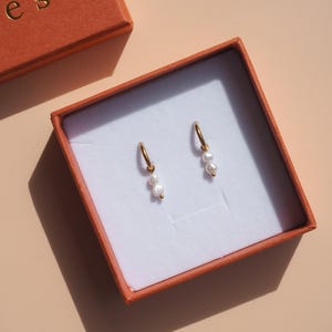 May include: Gold hoop earrings with three small, white pearls dangling from each hoop. The earrings are displayed in an orange box with a white interior. The box is open, revealing the jewelry. The background is a light peach color.