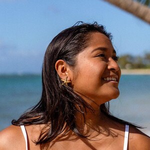 Puede incluir: Pendientes dorados de estrella de mar. La persona lleva los pendientes, tiene el pelo oscuro y una camiseta sin mangas blanca. El fondo incluye una playa, el océano y una palmera.