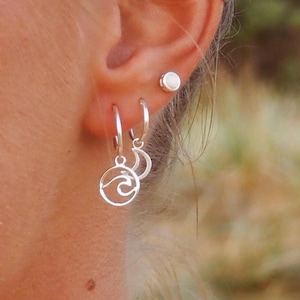 May include: A close-up of an ear with three pieces of jewelry. There is a small, round, white stud earring, and two silver hoop earrings. One hoop earring has a wave design, and the other has a crescent moon design.