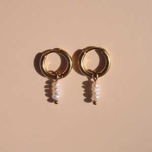 May include: A pair of gold-colored hoop earrings with a dangling cluster of three white pearls. The earrings are displayed against a light peach background, with shadows cast from the sunlight.