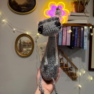 May include: A handmade gray crochet creature with large black eyes and a small head. The creature has a long neck and dangling legs. The plush toy is held in a person's hand. The background includes a neon flower light and a shelf of books.