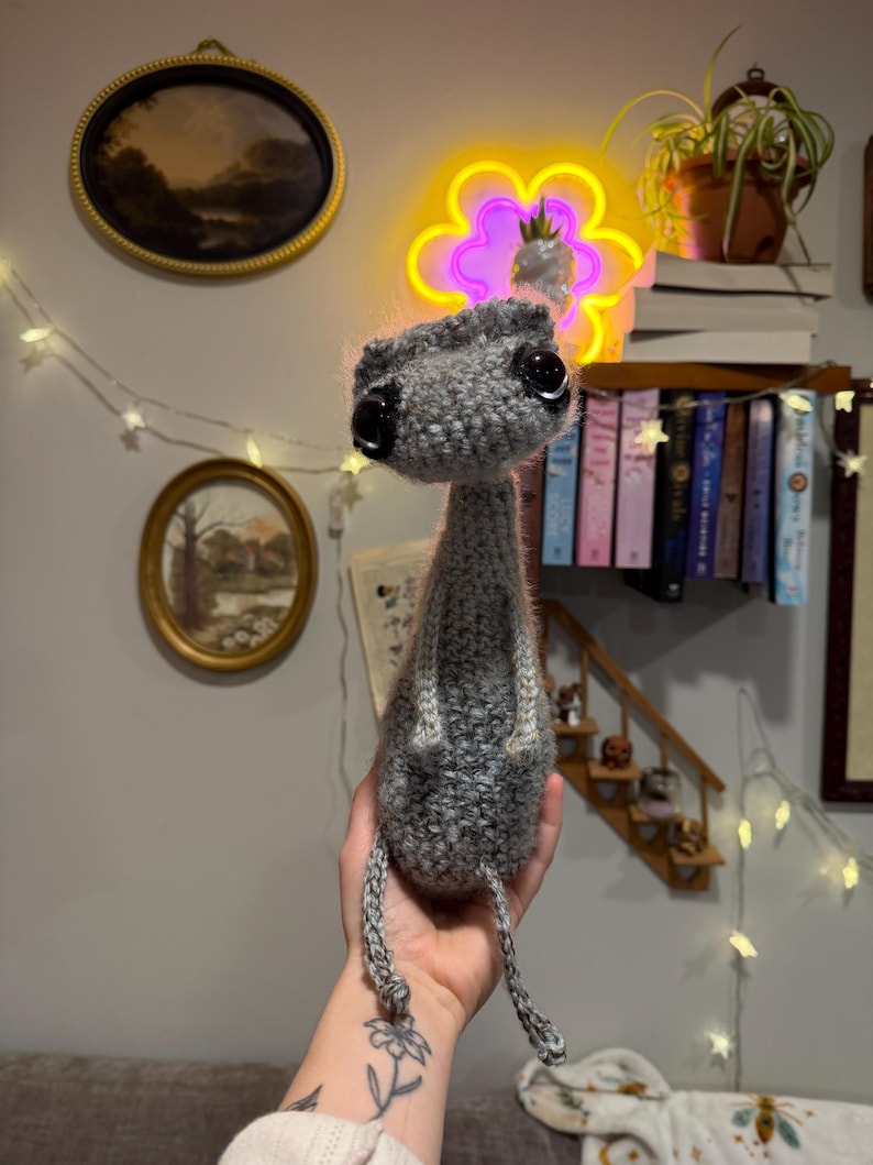 May include: A handmade gray crochet creature with large black eyes and a small head. The creature has a long neck and dangling legs. The plush toy is held in a person's hand. The background includes a neon flower light and a shelf of books.