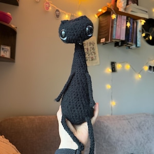 Może przedstawiać: Ręcznie robiona czarna szydełkowa lalka z dużymi, wyrazistymi oczami. Lalka ma smukłe ciało, małe ramiona i długie nogi. Tło przedstawia pokój z lampkami i neonem, tworzącym kapryśną atmosferę.