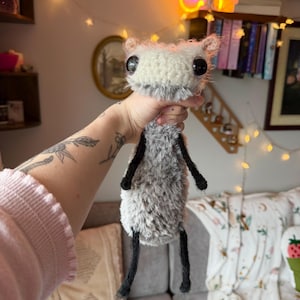 May include: A handmade plush creature with a fluffy white and grey body, large black eyes, and black limbs. The whimsical design features a soft, textured appearance, ideal for a unique gift or home decor item.