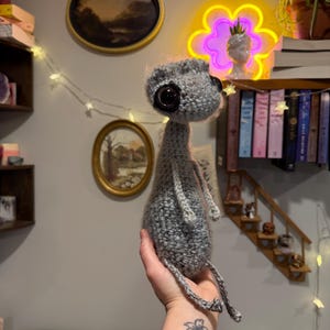May include: A handmade gray crocheted doll with large black button eyes and a small head. The doll has a slender body and long, dangling legs. The doll is held in a hand.