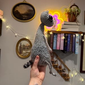 May include: A handmade gray crocheted alien doll with a large black eye and small legs and arms. The alien has a long neck and a small tail with black and gray stripes. The doll is held up in front of a wall with a neon flower light.