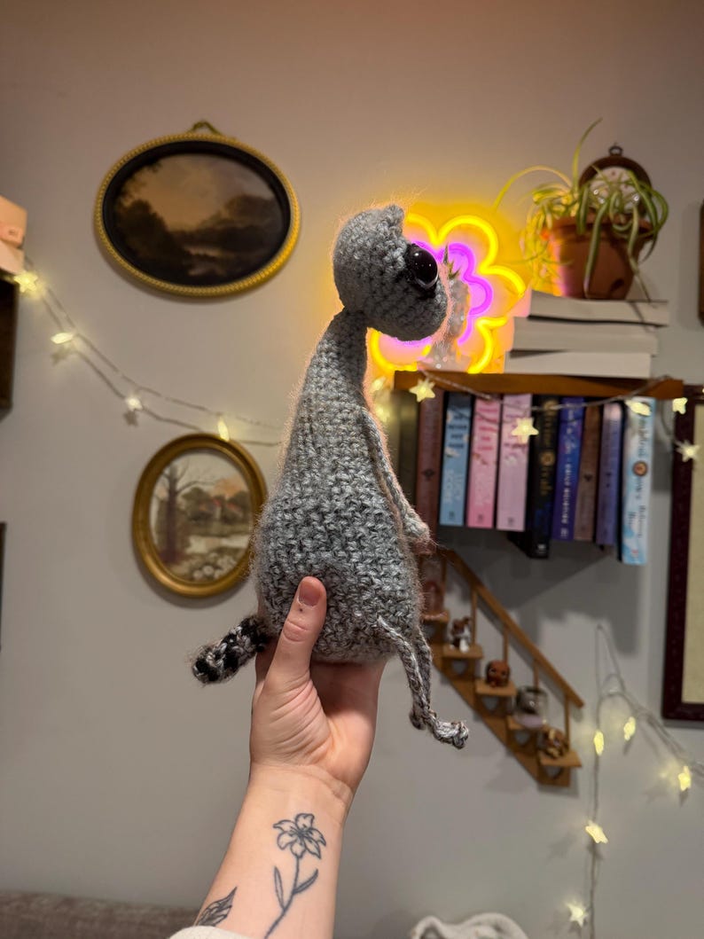 May include: A handmade gray crocheted alien doll with a large black eye and small legs and arms. The alien has a long neck and a small tail with black and gray stripes. The doll is held up in front of a wall with a neon flower light.
