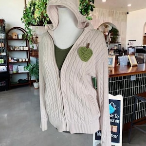Może przedstawiać: Jasny beżowy sweter z kapturem i zamkiem błyskawicznym, z zieloną naszywką w kształcie jabłka. Sweter jest prezentowany na manekinie, w połączeniu z zielonym topem. Tło obejmuje kawiarnię z roślinami i tablicą.