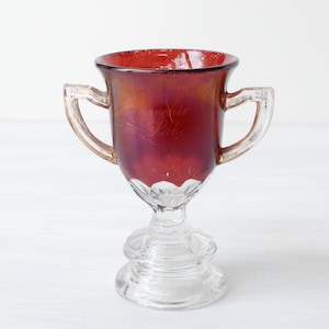 May include: A small, clear glass trophy with two handles and a red glass cup. The cup has a textured design and a gold iridescent sheen.