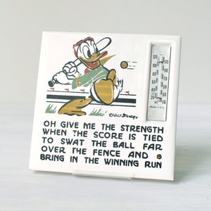 Könnte beinhalten: Weißer Keramik-Thermometer mit einem Cartoon-Bild von Donald Duck, der Baseball spielt. Das Thermometer hat ein schwarz-weißes Baseballfeld mit einem rot-weißen Baseball. Das Thermometer hat ein schwarz-weißes Thermometer mit einem roten Zeiger. Das Thermometer hat einen schwarz-weißen Text, der lautet: "OH GIVE ME THE STRENGTH WHEN THE SCORE IS TIED TO SWAT THE BALL FAR OVER THE FENCE AND BRING IN THE WINNING RUN"