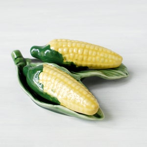 May include: Two ceramic corn on the cob salt and pepper shakers on a green leaf-shaped dish. The shakers are yellow with green tops and are sitting on a white surface.