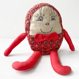 May include: A vintage handmade plush doll with an egg-shaped body. The doll features a red patterned fabric body, red arms and legs, a beige face with embroidered eyes and a smile, and a red bow tie. The doll is a whimsical toy.