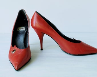 Puede incluir: Un par de zapatos de tacón alto de cuero rojo con punta puntiaguda. Los zapatos tienen un pequeño acento metálico en la parte delantera.