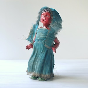 Può includere: Una bambola vintage con il viso rosso e un vestito blu. La bambola indossa un foulard blu e una collana d'oro. La bambola è fatta di legno dipinto e ha un'espressione fantasiosa.