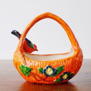 May include: A ceramic basket with an orange exterior and a handle. A red and black bird perches on the side. The basket is decorated with green leaves and blue and yellow flowers. The basket is a decorative item.