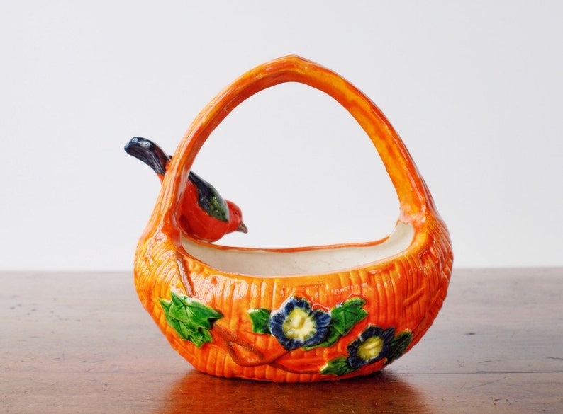May include: A ceramic basket with an orange exterior and a handle. A red and black bird perches on the side. The basket is decorated with green leaves and blue and yellow flowers. The basket is a decorative item.