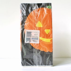 Puede incluir: Un paquete de servilletas de Halloween con fondo negro y una gran calabaza naranja con una cara de calabaza tallada. El paquete tiene una etiqueta que dice "Servilletas de cena de temporada Halloween", con un código de barras y un pequeño logotipo.