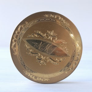 Op de afbeelding: Een goudkleurig metalen compact met een gegraveerd ontwerp met een gestileerd bloemmotief en de initialen "N.M.G."