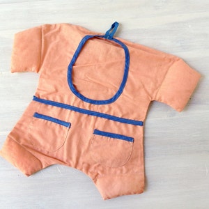 May include: A peach-coloured fabric baby onesie with blue trim around the neckline and pockets. The onesie has a loop at the top for hanging.