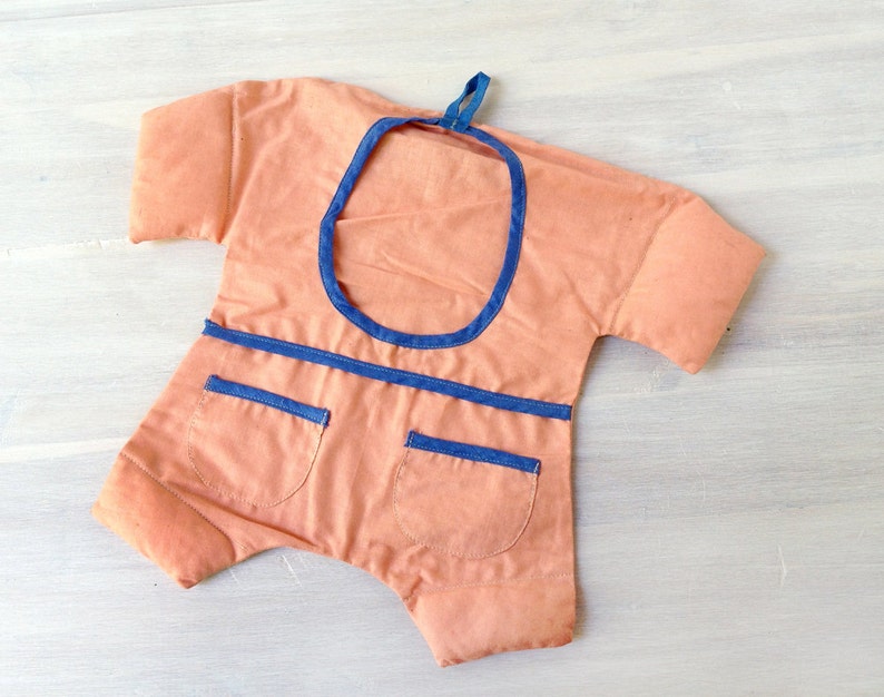 May include: A peach-coloured fabric baby onesie with blue trim around the neckline and pockets. The onesie has a loop at the top for hanging.