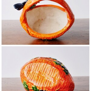 May include: A ceramic basket in orange, with a handle and a textured surface. The interior is white. It has green and blue floral details. The handle mirrors the basket's design. Measurements are in centimetres.