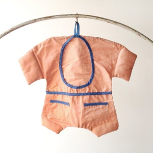 May include: A peach-coloured baby onesie with blue trim and two pockets. The onesie is hanging on a metal hanger.