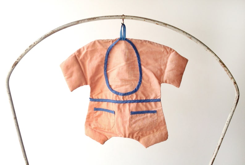 May include: A peach-coloured baby onesie with blue trim and two pockets. The onesie is hanging on a metal hanger.