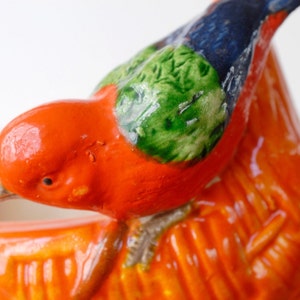 May include: Close-up of a ceramic bird figurine. The bird is mainly orange with green and blue accents on its wings and head. The figurine has a glossy finish and detailed textures.