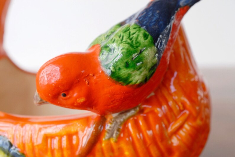 May include: Close-up of a ceramic bird figurine. The bird is mainly orange with green and blue accents on its wings and head. The figurine has a glossy finish and detailed textures.