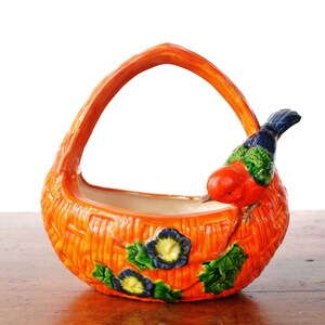 May include: Orange ceramic basket with a handle, featuring a textured woven design. A colourful bird with red, blue, and green plumage perches on the side. Blue and green floral accents decorate the basket's front.