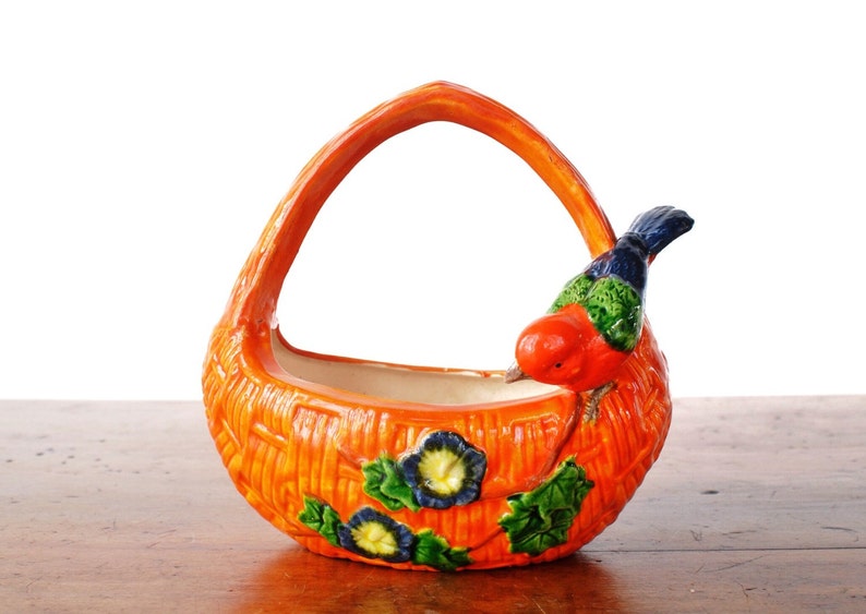 May include: Orange ceramic basket with a handle, featuring a textured woven design. A colourful bird with red, blue, and green plumage perches on the side. Blue and green floral accents decorate the basket's front.