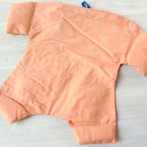 May include: A peach-coloured fabric baby doll with a blue ribbon tied around the neck. The doll has a simple design and is made of soft, plush fabric.