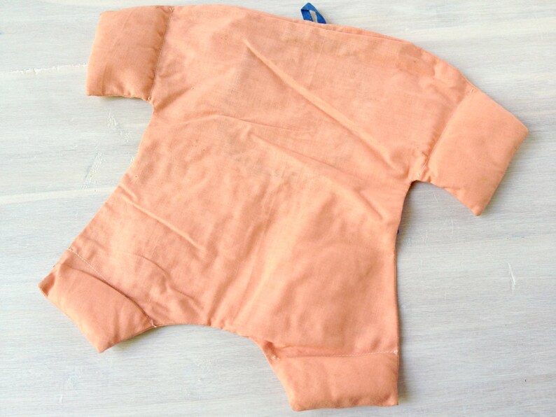 May include: A peach-coloured fabric baby doll with a blue ribbon tied around the neck. The doll has a simple design and is made of soft, plush fabric.