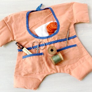 May include: A peach-coloured fabric sewing kit shaped like a doll with blue trim. The kit has pockets for storing sewing supplies, including scissors, thread, and a needle.