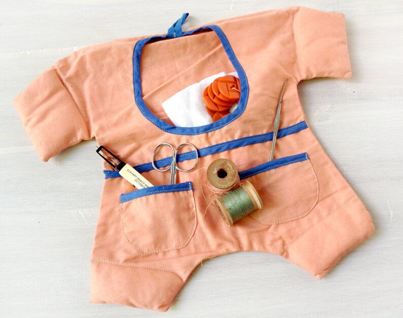 May include: A peach-coloured fabric sewing kit shaped like a doll with blue trim. The kit has pockets for storing sewing supplies, including scissors, thread, and a needle.
