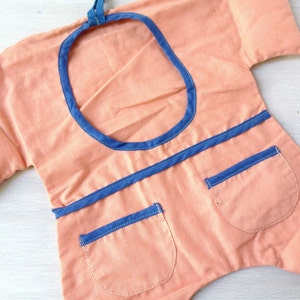 May include: A peach-coloured fabric garment with a blue trim around the neckline and pockets. The garment has a round opening at the top and two pockets on the front.