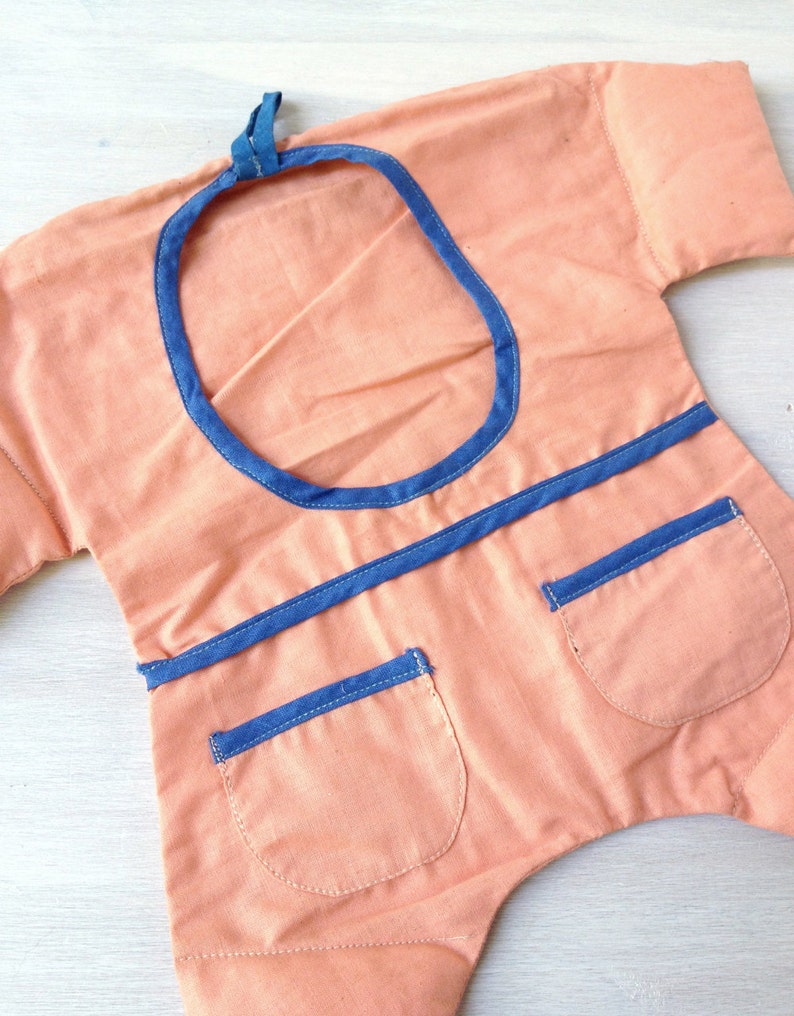 May include: A peach-coloured fabric garment with a blue trim around the neckline and pockets. The garment has a round opening at the top and two pockets on the front.