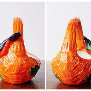 May include: Orange ceramic basket with a woven texture and a curved handle. The basket features a bird with red, green, and black details on the side. Green leaves are painted on the base of the basket.