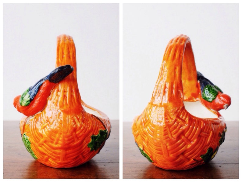 May include: Orange ceramic basket with a woven texture and a curved handle. The basket features a bird with red, green, and black details on the side. Green leaves are painted on the base of the basket.