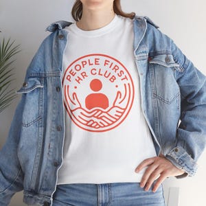 May include: White t-shirt with a red circular graphic that reads "PEOPLE FIRST HR CLUB." The graphic features a person icon and two hands. The person is wearing a light blue denim jacket and blue jeans.