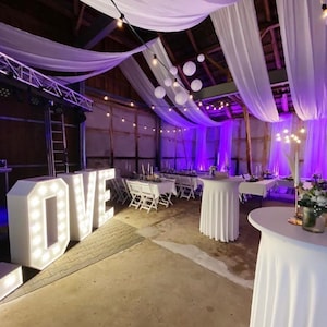 May include: An event venue decorated with white drapes, fairy lights, and purple uplighting. Large illuminated "LOVE" letters are in the foreground. Tables are set for a reception, creating a festive atmosphere.