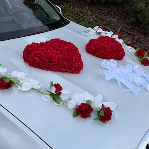 May include: White car hood decorated with red rose heart shapes and white floral garlands. The hood also features white bows and additional red roses, creating a festive and romantic display.
