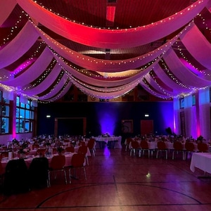 Peut inclure: Une grande salle décorée pour un événement. Des rideaux en tissu blanc pendent du plafond, avec des guirlandes lumineuses et un éclairage coloré. Les tables sont dressées avec des chaises rouges, créant une ambiance festive.