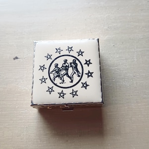 Op de afbeelding: Kleine, vierkante doos met scharnieren en een crèmekleurige bovenkant. De bovenkant heeft een zwarte illustratie van drie figuren in een cirkel, omringd door een ring van zwarte sterren. De doos heeft een zilverkleurig metalen frame.