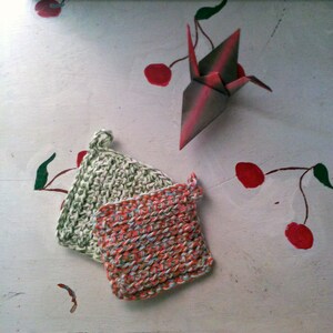 May include: Two crocheted dishcloths, one green and one multi-colored, are displayed on a white surface with a red origami crane and painted red cherries.